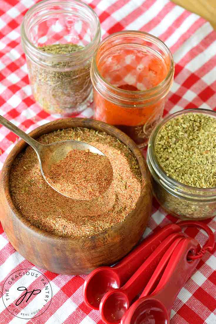 A wooden bowl filled with this Cajun Seasoning Recipe, sits with a serving spoon in the bowl and jars of spices and measuring spoons around it.