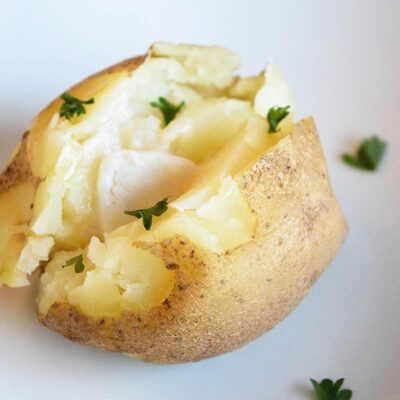 A single, delicious, potato sits on a white plate with a pat of butter inside the potato which is cut down the middle with some fresh parsley sprinkled over the top in this Instant Pot Baked Potatoes recipe.