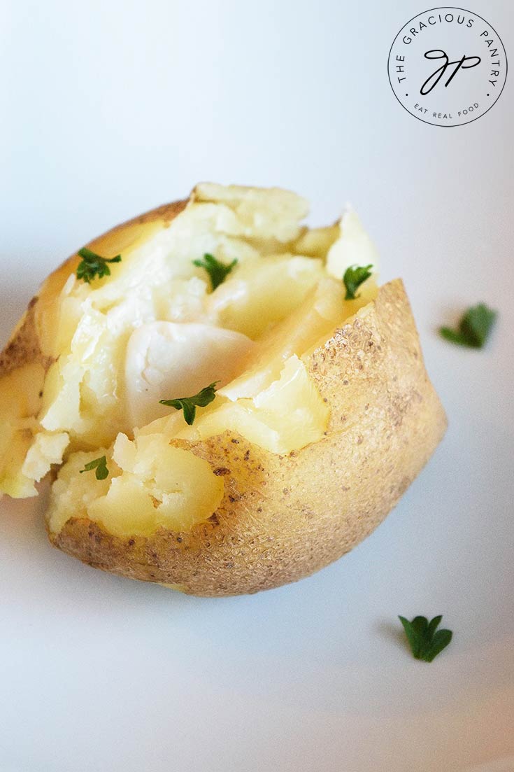 A single, delicious, potato sits on a white plate with a pat of butter inside the potato which is cut down the middle with some fresh parsley sprinkled over the top in this Instant Pot Baked Potatoes recipe.