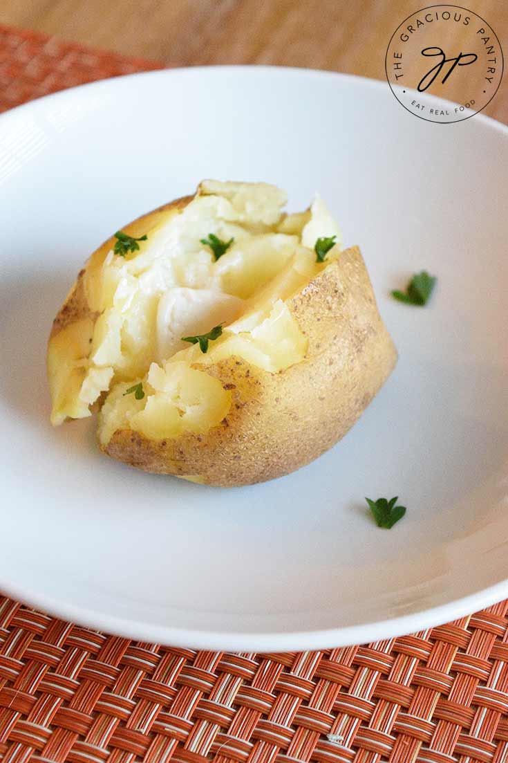 A single potato on a white plate, sits on a table, ready to enjoy with melted butter and a sprinkle of fresh, chopped parsley over the top.