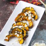 A platter of this Roasted Acorn Squash With Feta sits on a table with a bowl of feta crumbles next to it, all ready to serve.
