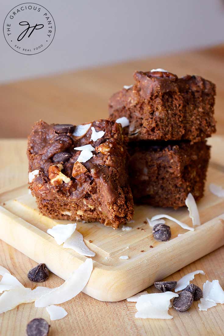 A stack of German Chocolate Brownies sits on a cutting board with coconut flakes and chocolate chips strewn around the board.