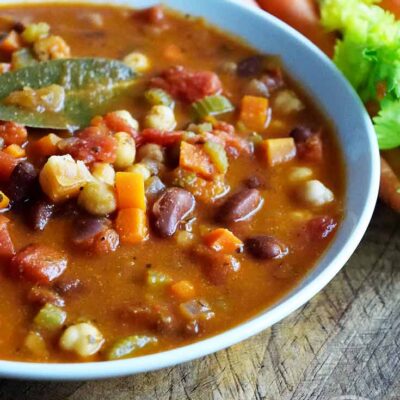 A white bowl filled with this Minestrone Soup sits ready to enjoy. Carrots and celery sit off in the distance.