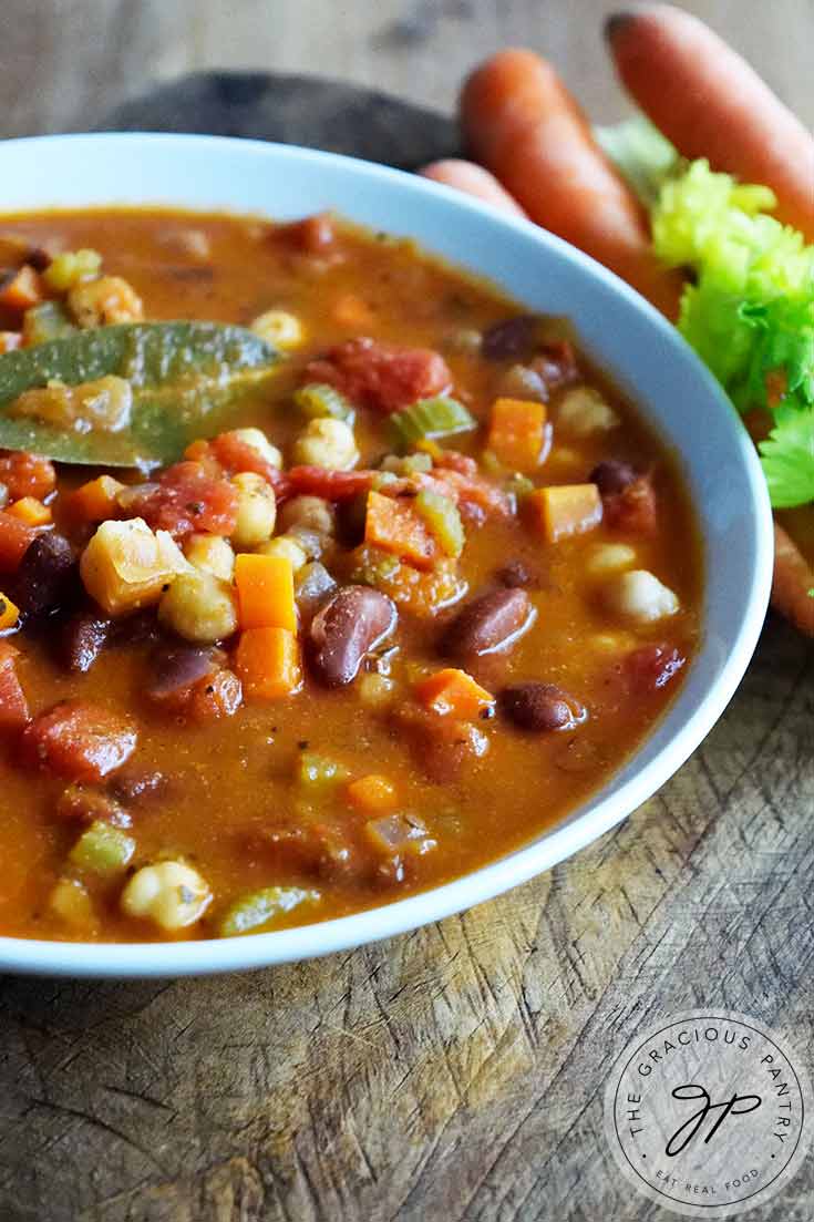 A white bowl filled with this Minestrone Soup sits ready to enjoy. Carrots and celery sit off in the distance.