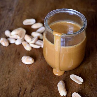 A jar of this peanut sauce sits with a drip coming down the side of the jar onto the table. A few peanuts are scattered around the jar.