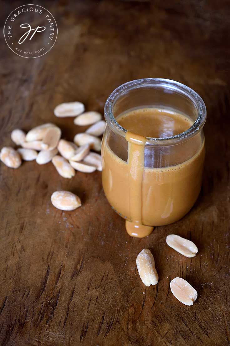 A jar of this peanut sauce sits with a drip coming down the side of the jar onto the table. A few peanuts are scattered around the jar.