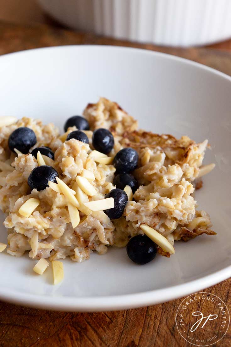 A portion of baked oatmeal sits in a white bowl, topped with fresh blueberries.