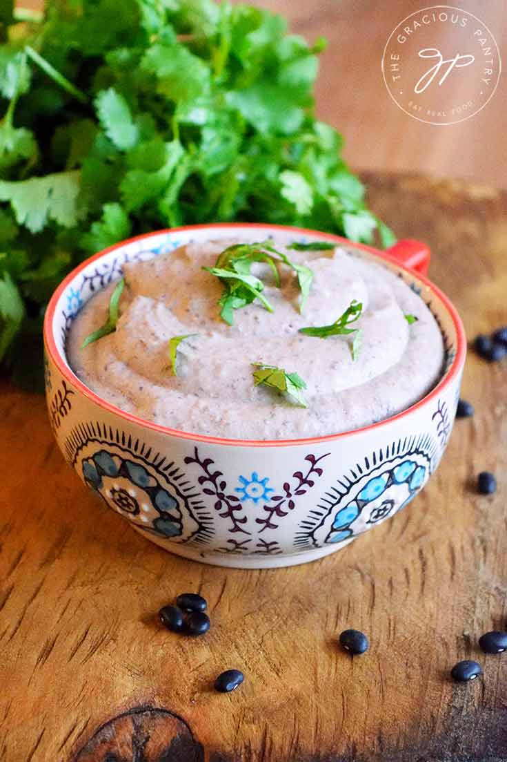 A decorative mug sits filled with this Black Bean Hummus. Fresh cilantro is sprinkled over the top and a fresh bunch of cilantro is laying behind the mug.