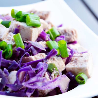A white bowl sits on a table filled with purple cabbage, green onions and chunks of tofu in this Tofu Stir Fry Recipe.