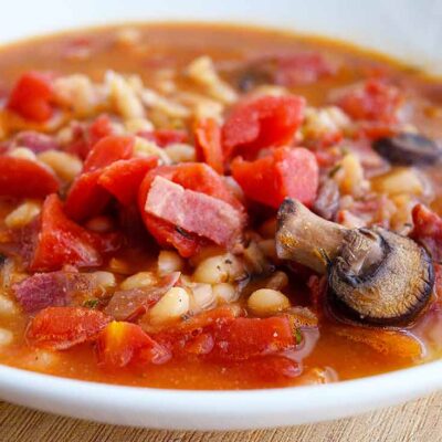 ThisItalian Navy Bean Soup fills a white bowl. You can see bits of bacon, mushrooms, tomatoes and plenty of navy beans sitting in a delicious broth.