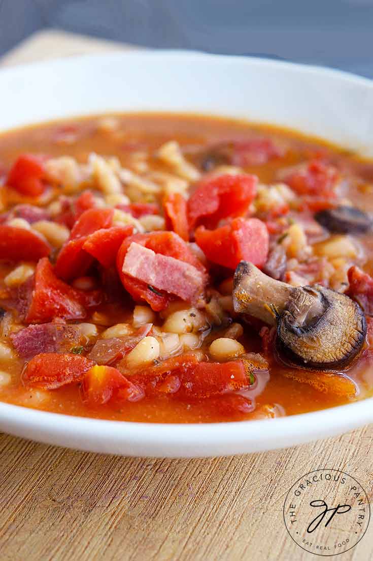 ThisItalian Navy Bean Soup fills a white bowl. You can see bits of bacon, mushrooms, tomatoes and plenty of navy beans sitting in a delicious broth.