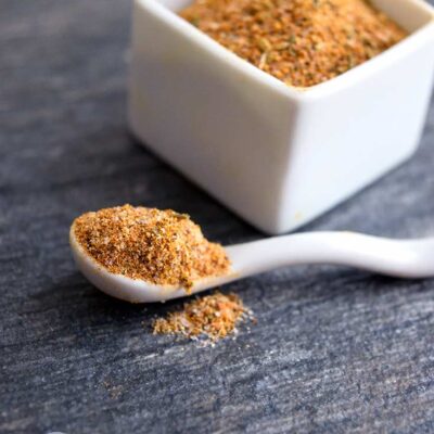 A white spice container sits filled with this adobo seasoning recipe. A spoon full of the seasoning sits in front of it on the table.