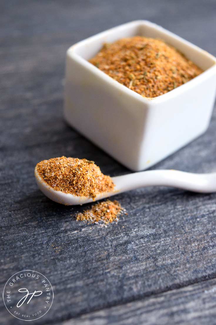 A white spice container sits filled with this adobo seasoning recipe. A spoon full of the seasoning sits in front of it on the table.