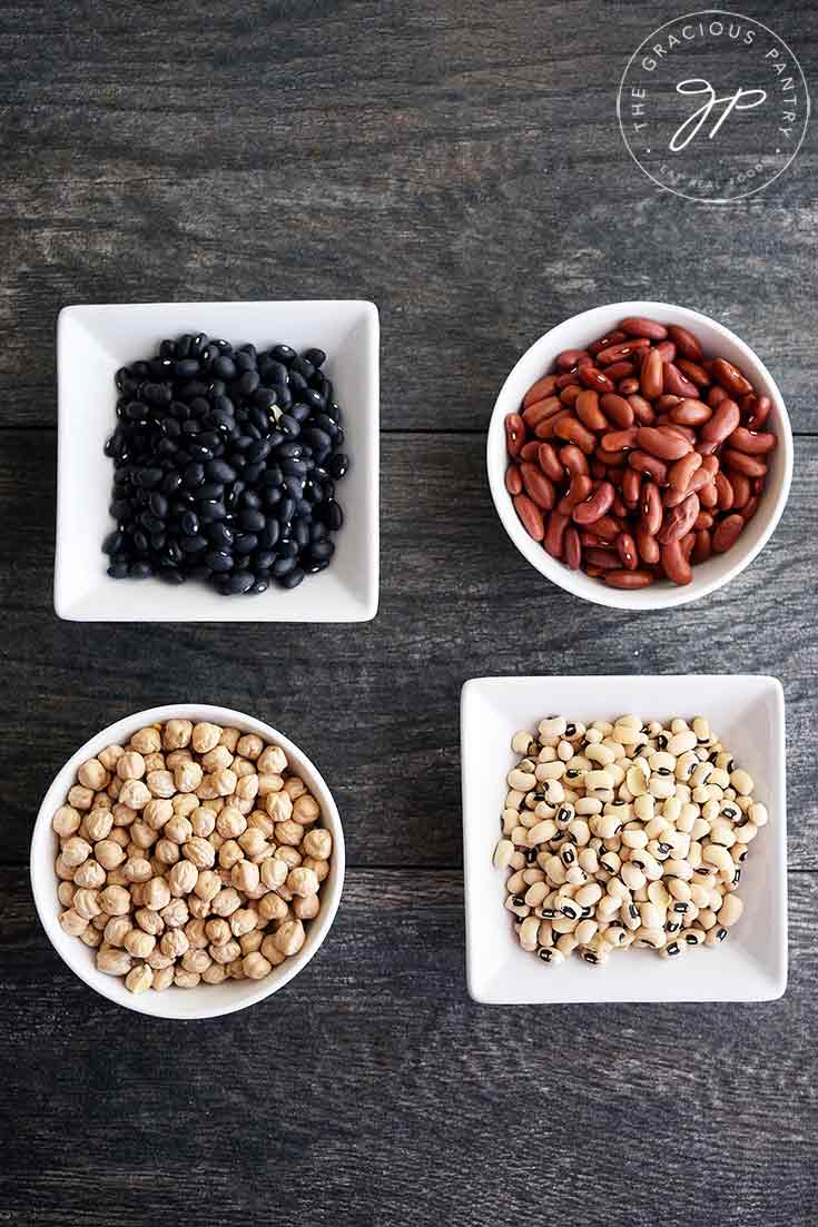 An overhead view of four different types of beans in separate, white bowls in this guide to How To Cook Dry Beans.