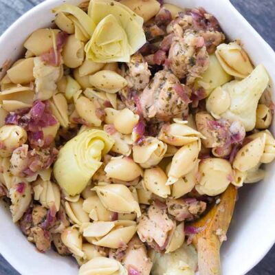 An overhead view looking down into a serving bowl of this delicious artichoke chicken pasta. A wooden spoon is in the bowl, ready to serve the pasta.