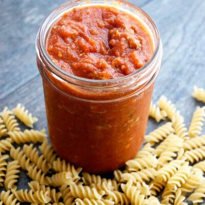 A jar of homemade spaghetti sauce sits with the lid off and some dry spiral pasta lays at the base of the jar.