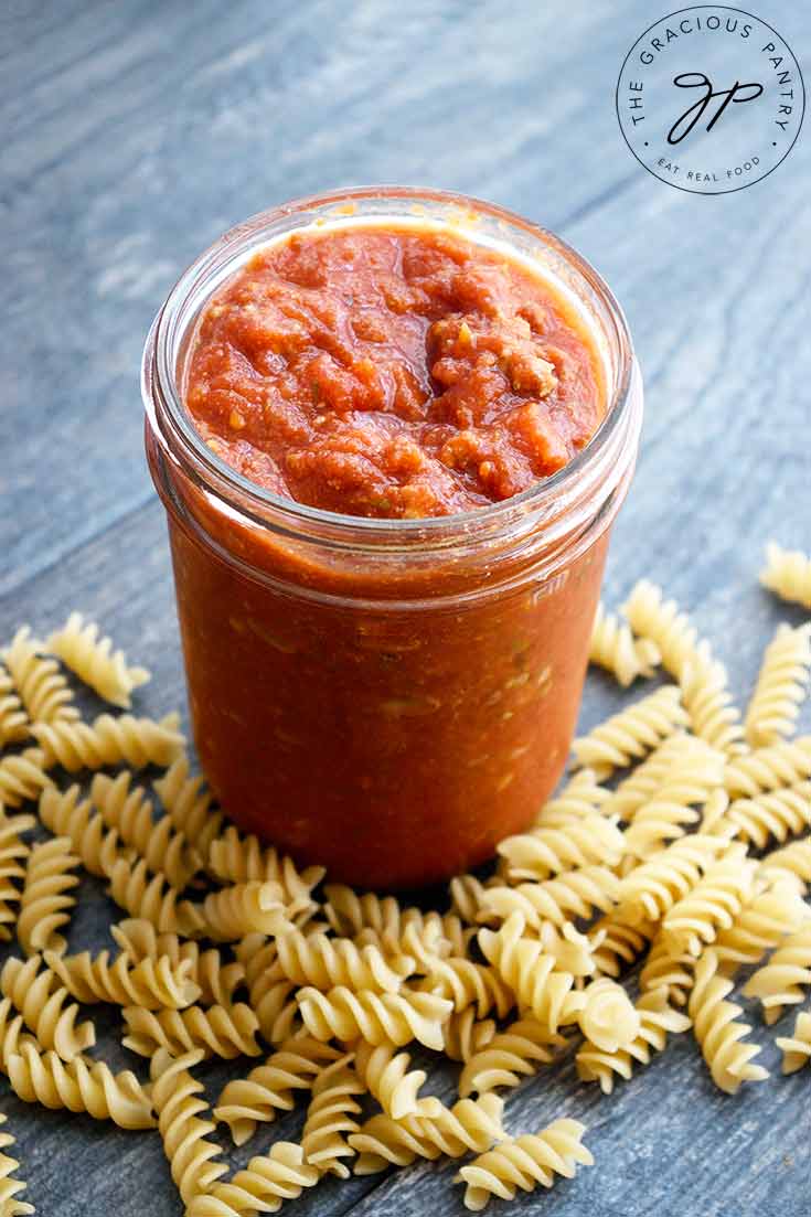 A jar of homemade spaghetti sauce sits with the lid off and some dry spiral pasta lays at the base of the jar.