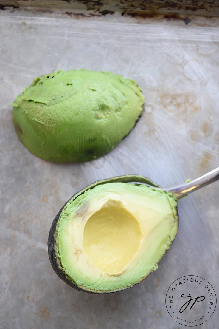 An avocado half with the skin removed, sits on a pan, ready to freeze. A spoon sits between the skin and flesh of the other half.