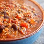 A blue bowl filled with this Quinoa Chili sits ready to eat. You can see the quinoa and black beans with bits of tomatoes.