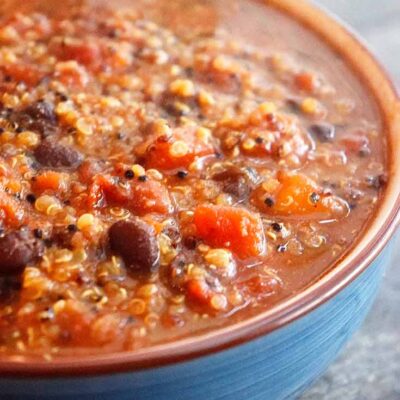 A blue bowl filled with this Quinoa Chili sits ready to eat. You can see the quinoa and black beans with bits of tomatoes.