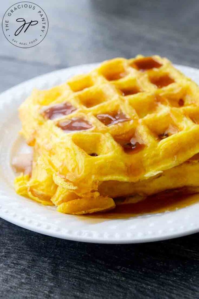 A short stack of egg waffles sit on a white plate, topped with maple syrup.