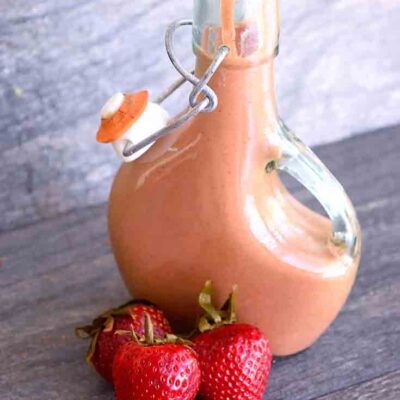 A bottle of this Strawberry Balsamic Vinaigrette sits on a table with three fresh strawberries laying in front of it.