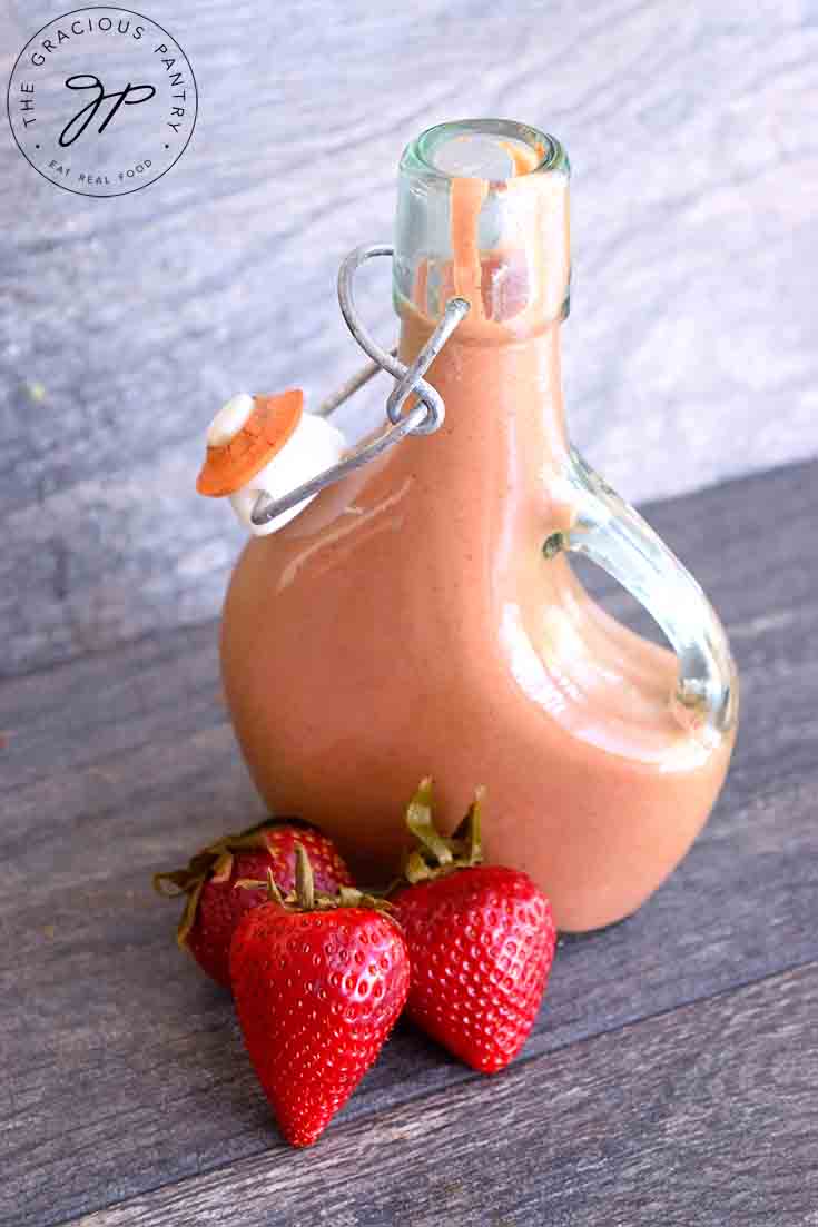 A bottle of this Strawberry Balsamic Vinaigrette sits on a table with three fresh strawberries laying in front of it.