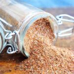 A spice jar filled with this Chili Spice Recipe sits tipped over with the spices spilling out onto a wood surface.