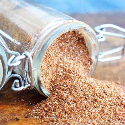 A spice jar filled with this Chili Spice Recipe sits tipped over with the spices spilling out onto a wood surface.