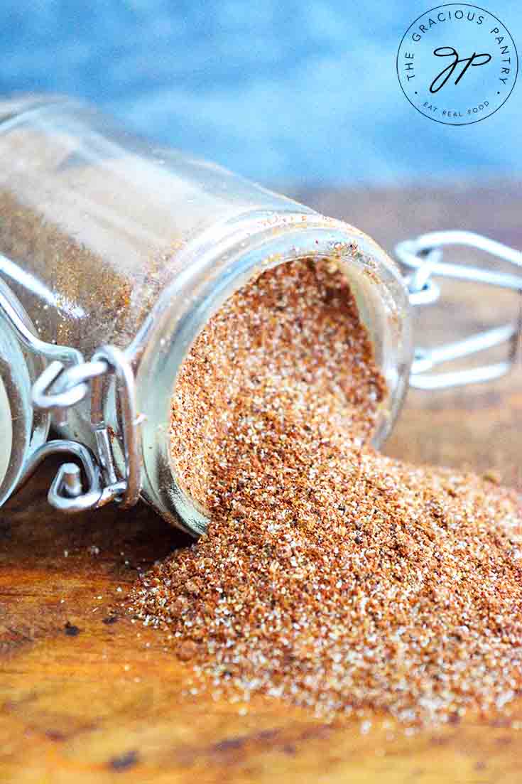 A spice jar filled with this Chili Seasoning Recipe sits tipped over with the spices spilling out onto a wood surface.