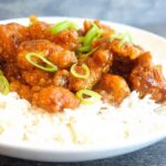 This Easy Orange Chicken sits on a bed of rice, served in a white bowl and topped with sliced, green onions.