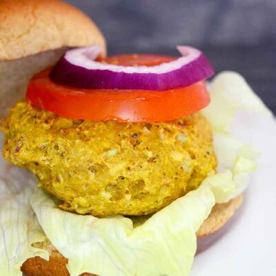 A Tuna Burger sits on a white plate topped with a slice of tomato and a slice of red onion, atop a whole grain burger bun.