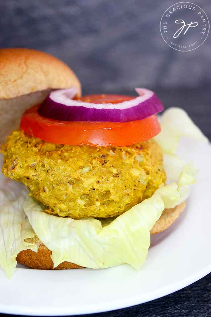 One of four Tuna Burgers sits on a white plate topped with a slice of tomato and a slice of red onion, atop a whole grain burger bun.