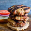 A stack of three Grilled Turkey Burgers sit on a whole grain but with the other half behind it loaded with lettuce, tomato and onion slices.