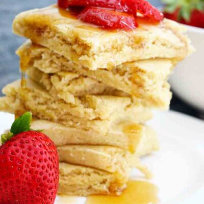 A stack of square, sheet pan pancakes sits piled high with strawberry slices and maple syrup on top.