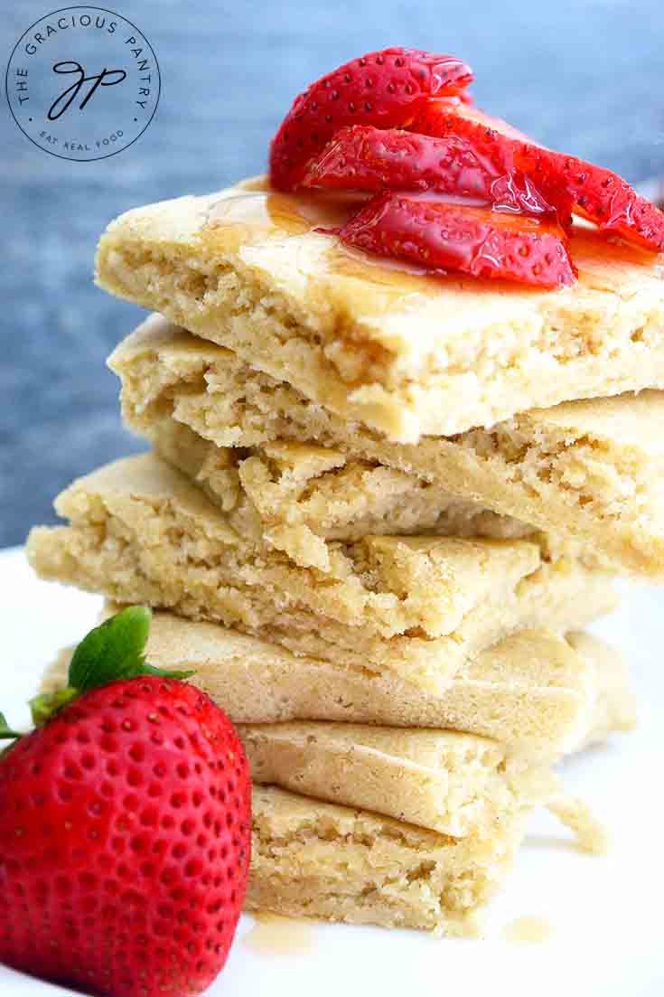 And up close view of this stack of Sheet Pan Pancakes topped with strawberries and maple syrup. A whole berry sits on the plate next to the stack of pancakes.