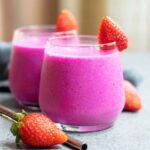 Two glasses of bright pink dragon fruit smoothie sit on a table with strawberries.
