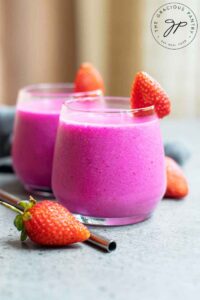 Two glasses of bright pink dragon fruit smoothie sit on a table with strawberries.