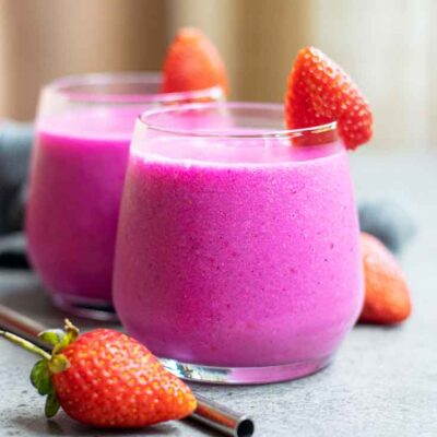 Two glasses of bright pink dragon fruit smoothie sit on a table with strawberries.