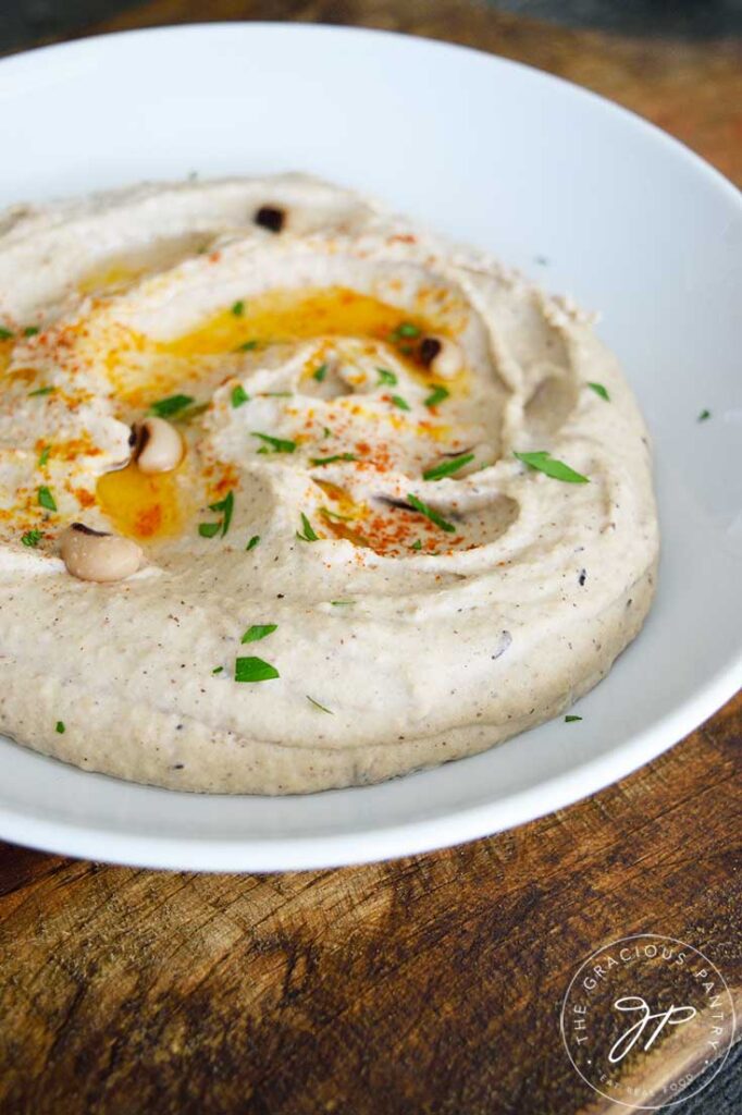 A side view of a white bowl with Black Eyed Pea Hummus, topped with oil, paprika and a bit of fresh herbs.