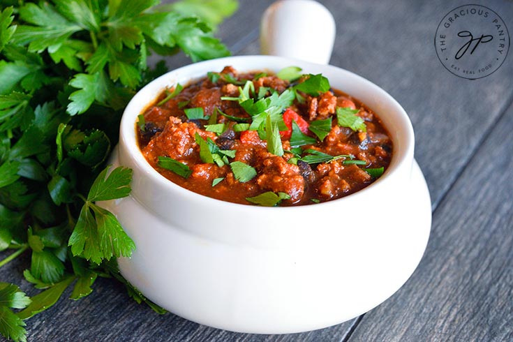 Horizontal shot of a white crock filled with turkey chili and garnished with chopped parsley.