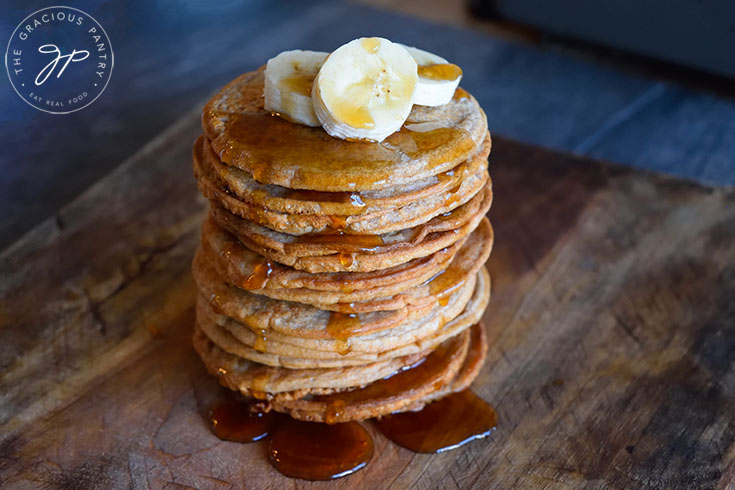 A horizontal side view of this Banana Pancakes Recipe. A stack of banana pancakes sits on a cutting board, topped with banana slices and maple syrup which is dripping down the sides onto the board.