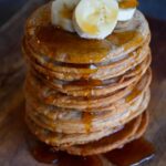 Banana Pancakes in a tall stack with three banana slices on top and maple syrup dripping down over the sides onto a wooden surface.