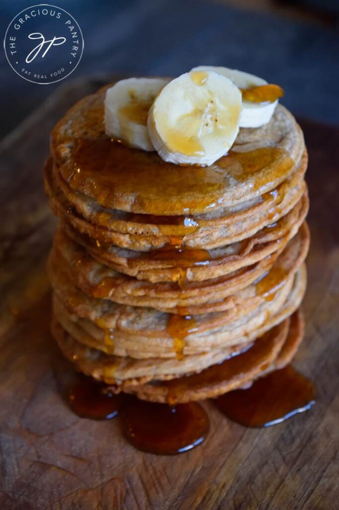 Banana Pancakes in a tall stack with three banana slices on top and maple syrup dripping down over the sides onto a wooden surface.