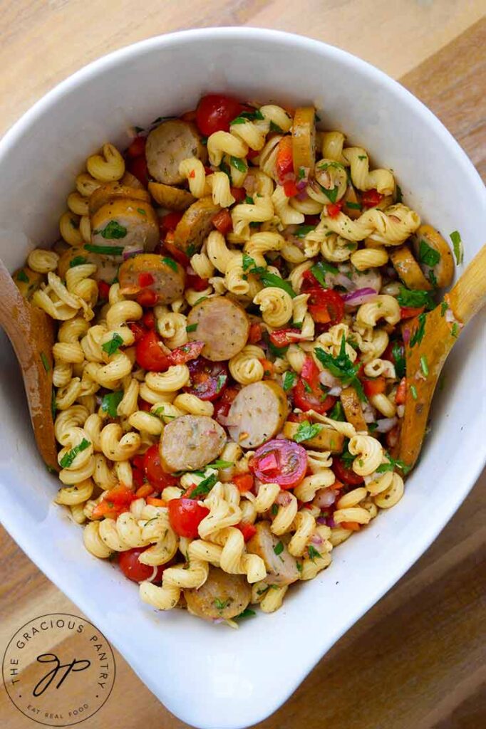 Overhead shot looking down into a large, white mixing bowl of cavatappi pasta with sausage slice, bell peppers, tomatoes and fresh herbs.