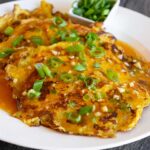 A plate of Egg Foo Young with a side of sliced green onions. Gravy slathered over the top with fresh green onions on top.