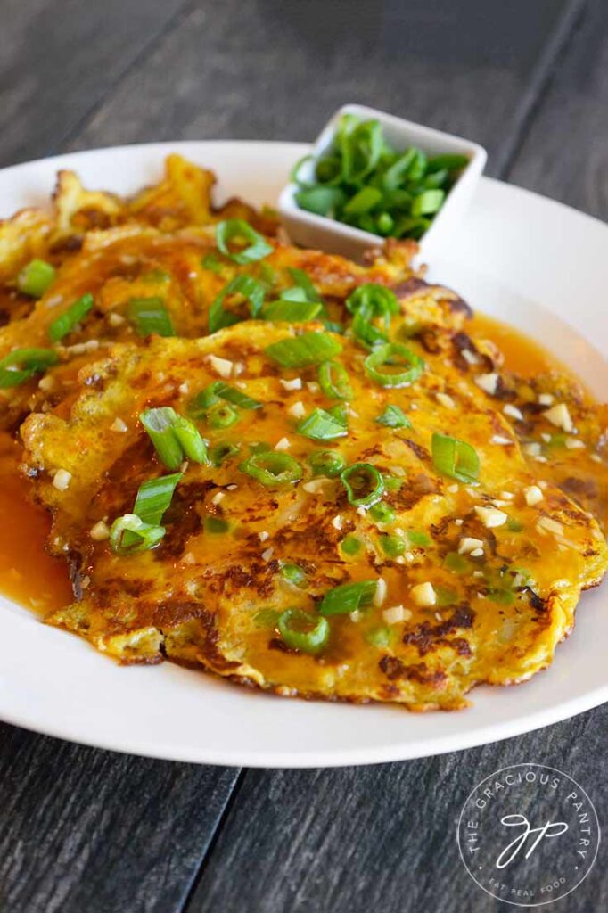 A plate of Egg Foo Young with a side of sliced green onions. Gravy slathered over the top with fresh green onions on top.