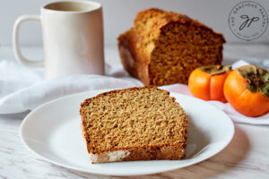 A plate with a slice of Persimmon Bread just cut from the loaf that sits behind it.
