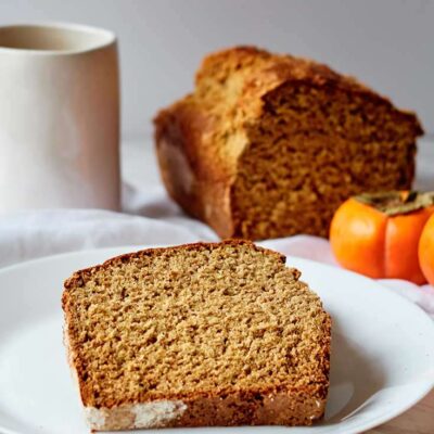 A slice of persimmon bread served on a plate. The cut loaf sits behind it and a mug sits at the back to the left.