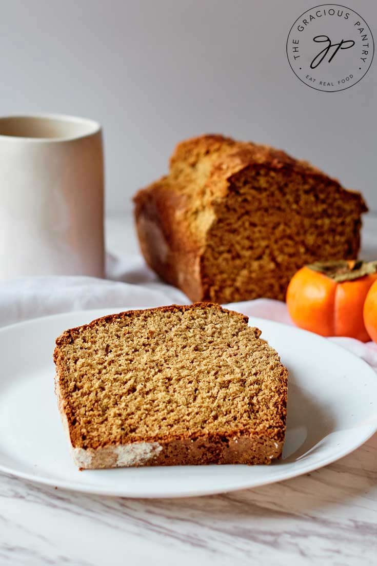 A slice of persimmon bread served on a plate. The cut loaf sits behind it and a mug sits at the back to the left.